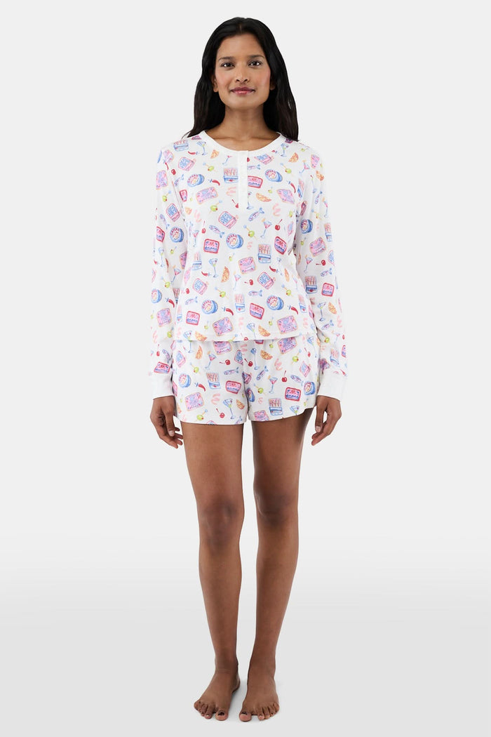 A person wears the Roller Rabbit Tinned or Twist Ashley Top, a white long-sleeve with colorful food and drink prints, while sitting on a couch with red and green patterned pillows in the background. The image is cropped at the shoulders.