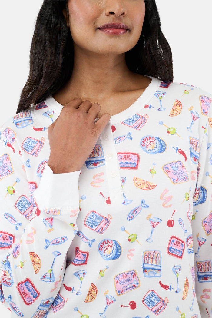 A person wears the Roller Rabbit Tinned or Twist Ashley Top, a white long-sleeve with colorful food and drink prints, while sitting on a couch with red and green patterned pillows in the background. The image is cropped at the shoulders.