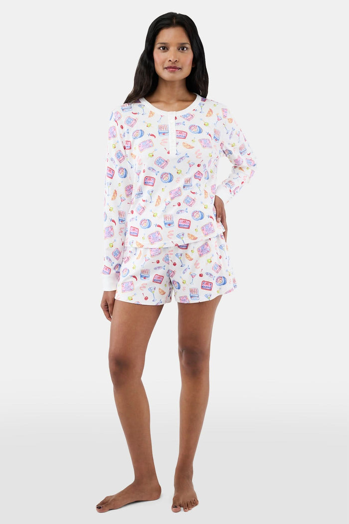 A person wears the Roller Rabbit Tinned or Twist Ashley Top, a white long-sleeve with colorful food and drink prints, while sitting on a couch with red and green patterned pillows in the background. The image is cropped at the shoulders.