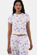 A woman with long dark hair wears the Roller Rabbit Tinned or Twist Baby Tee, featuring a vibrant foodie print with cocktails and snacks on a white background, paired with matching pants against a simple backdrop.