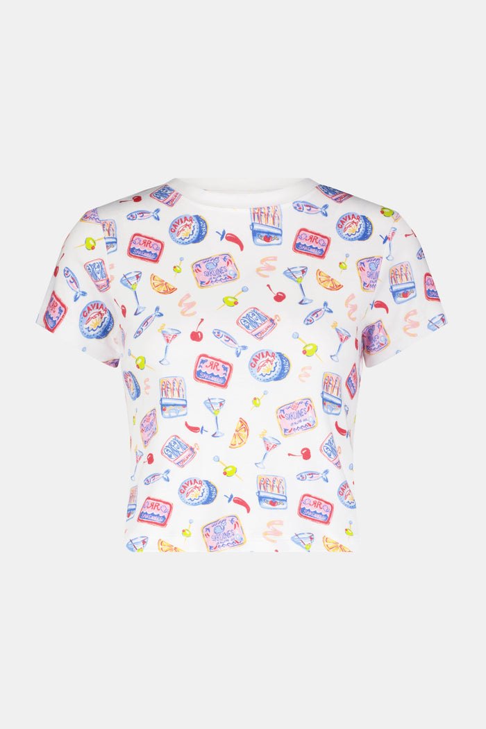 A woman with long dark hair wears the Roller Rabbit Tinned or Twist Baby Tee, featuring a vibrant foodie print with cocktails and snacks on a white background, paired with matching pants against a simple backdrop.