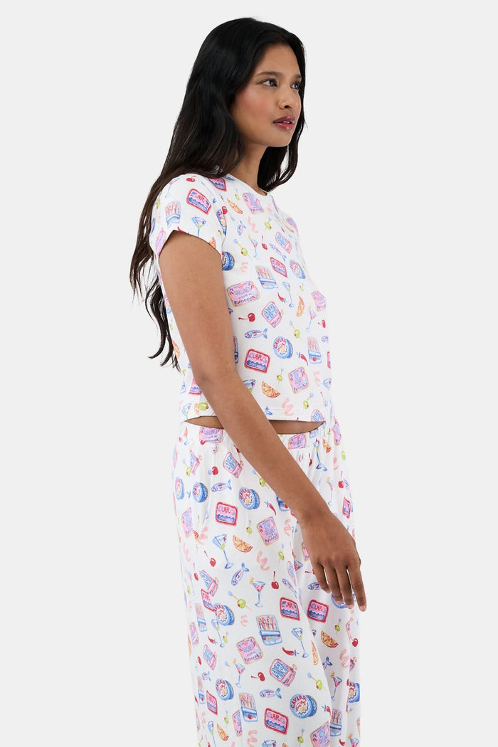 A woman with long dark hair wears the Roller Rabbit Tinned or Twist Baby Tee, featuring a vibrant foodie print with cocktails and snacks on a white background, paired with matching pants against a simple backdrop.