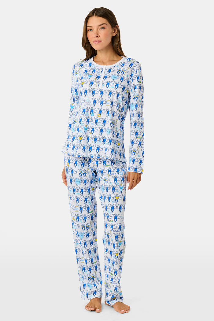 A woman with long brown hair sits on outdoor steps, gazing to the side while effortlessly showcasing her stylish look in Roller Rabbit’s Tri Delta Monkey Pajamas, paired with sunglasses and a navy jacket draped over her shoulders.