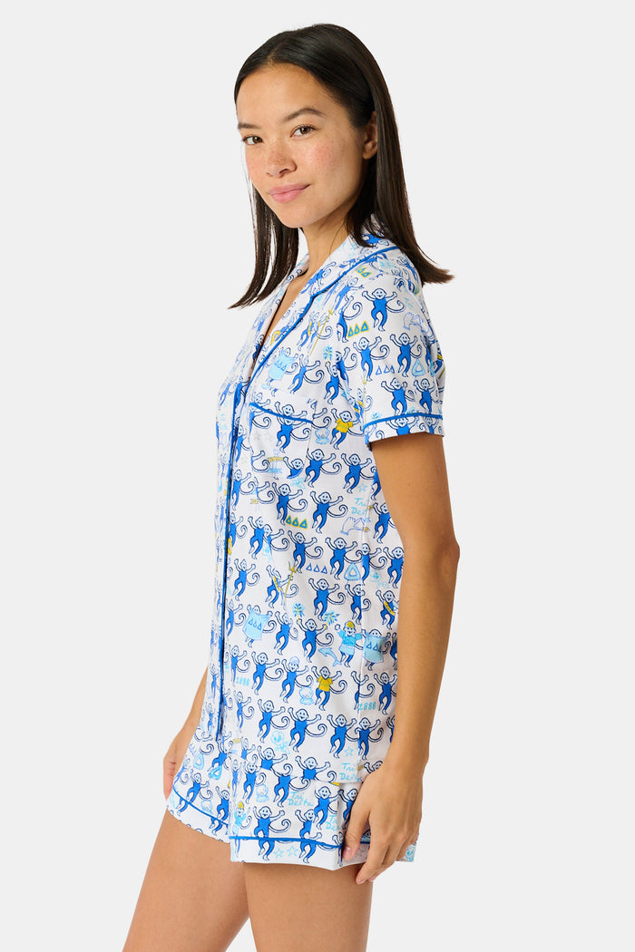 A woman wearing blue and white patterned Tri Delta Monkey Polo Pajamas by Roller Rabbit holds a large striped bag. She stands outdoors near a brick wall and green plants, with sunlight filtering through the leaves.