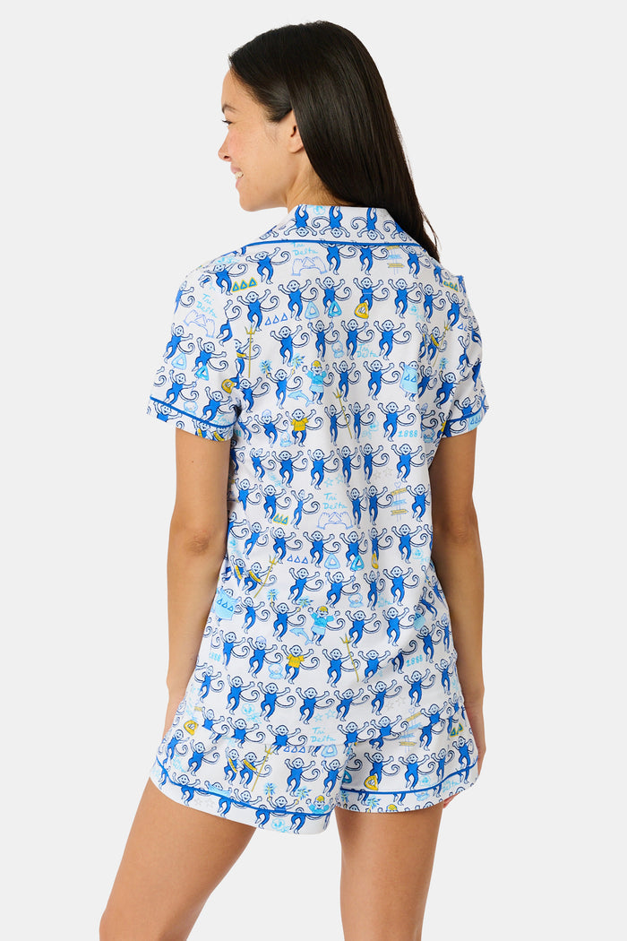 A woman wearing blue and white patterned Tri Delta Monkey Polo Pajamas by Roller Rabbit holds a large striped bag. She stands outdoors near a brick wall and green plants, with sunlight filtering through the leaves.