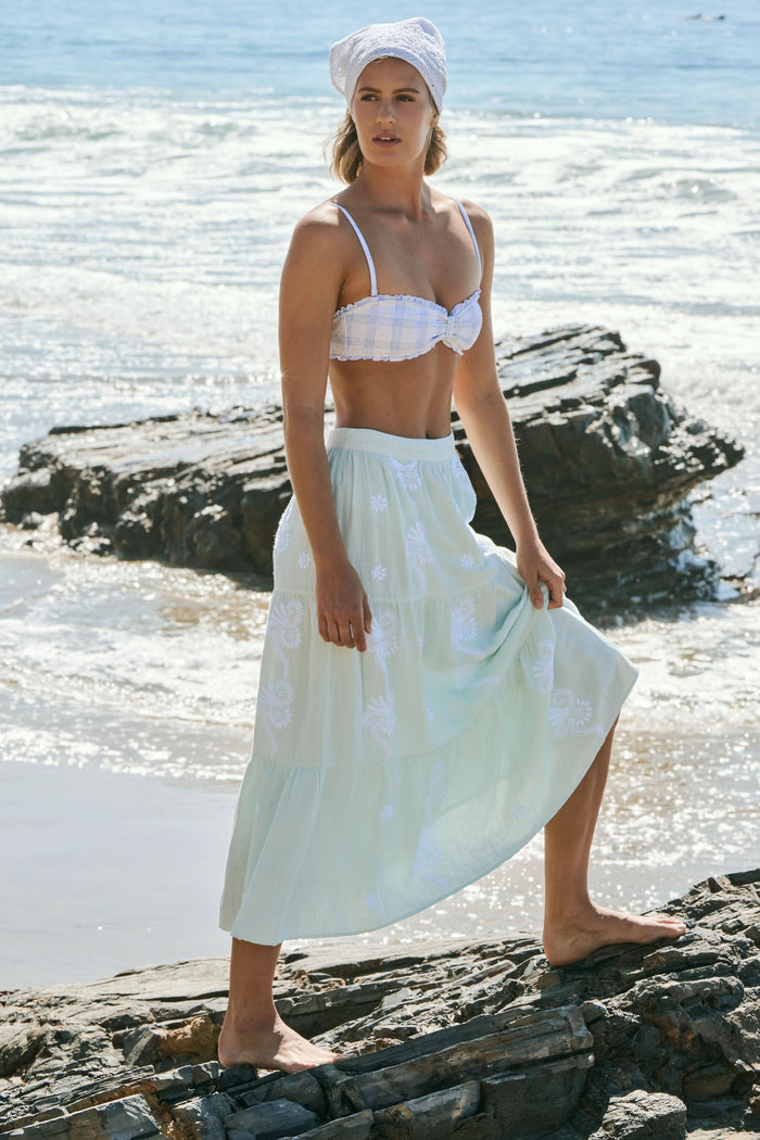A woman stands barefoot on rocks by the ocean, wearing a white headscarf, a plaid white bikini top, and the light green Vanna Embroidery Carlisa Skirt by Roller Rabbit. Gentle waves and sunlight shine in the background.