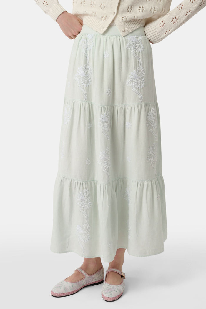 A woman stands barefoot on rocks by the ocean, wearing a white headscarf, a plaid white bikini top, and the light green Vanna Embroidery Carlisa Skirt by Roller Rabbit. Gentle waves and sunlight shine in the background.