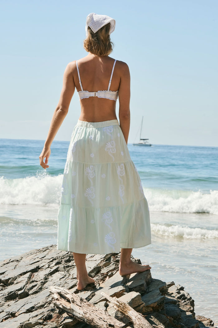 A woman stands barefoot on rocks by the ocean, wearing a white headscarf, a plaid white bikini top, and the light green Vanna Embroidery Carlisa Skirt by Roller Rabbit. Gentle waves and sunlight shine in the background.