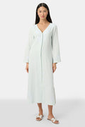 A woman faces forward wearing the Roller Rabbit Vanna Embroidery Rachel Caftan—a long, light green linen dress with subtle white embroidery and long sleeves—paired with white sandals against a plain light gray background.