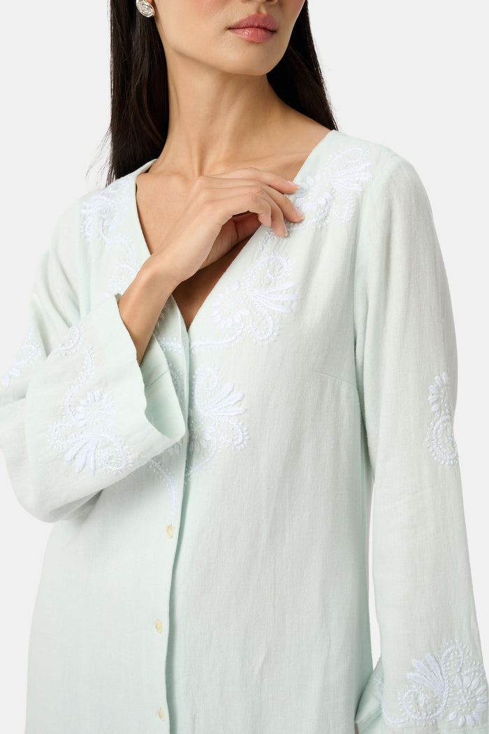 A woman faces forward wearing the Roller Rabbit Vanna Embroidery Rachel Caftan—a long, light green linen dress with subtle white embroidery and long sleeves—paired with white sandals against a plain light gray background.