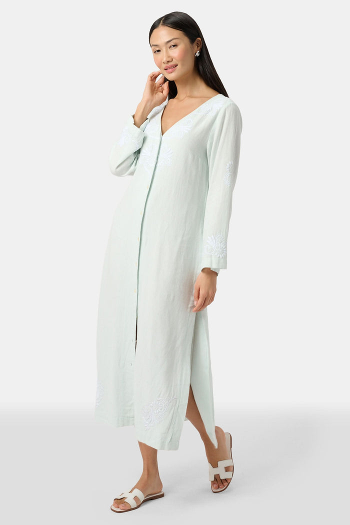 A woman faces forward wearing the Roller Rabbit Vanna Embroidery Rachel Caftan—a long, light green linen dress with subtle white embroidery and long sleeves—paired with white sandals against a plain light gray background.