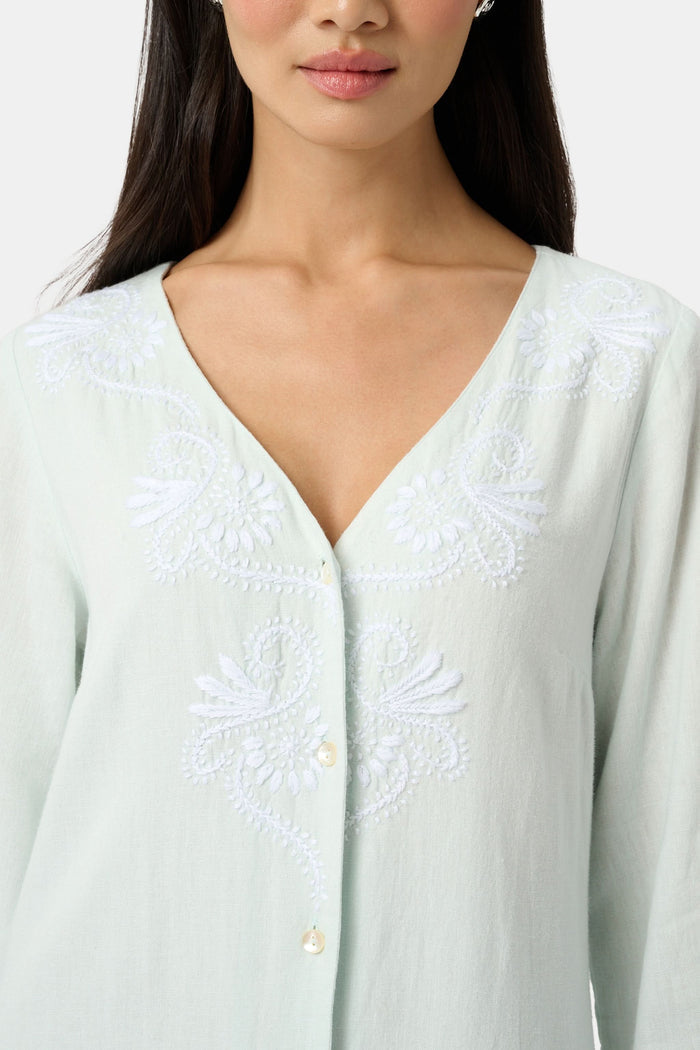 A woman faces forward wearing the Roller Rabbit Vanna Embroidery Rachel Caftan—a long, light green linen dress with subtle white embroidery and long sleeves—paired with white sandals against a plain light gray background.