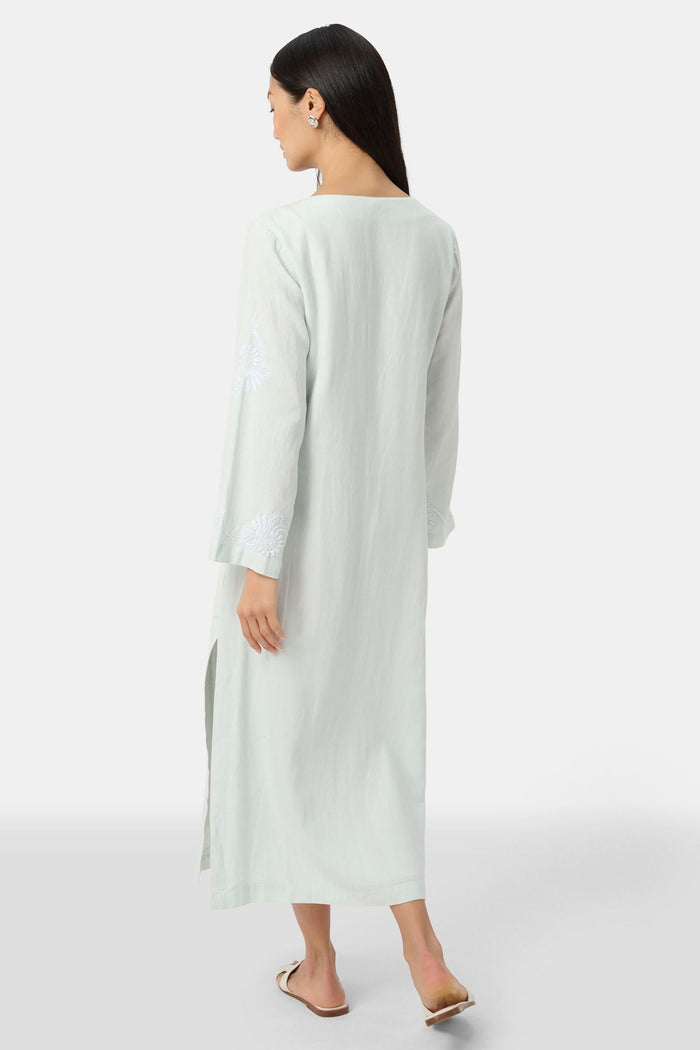 A woman faces forward wearing the Roller Rabbit Vanna Embroidery Rachel Caftan—a long, light green linen dress with subtle white embroidery and long sleeves—paired with white sandals against a plain light gray background.