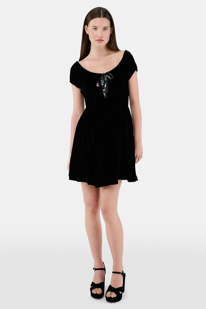A woman faces forward in the Roller Rabbit Velvet Tilly Dress, a short-sleeve black velvet mini with a floral bow at the neckline, paired with black platform sandals, set against a plain light background.