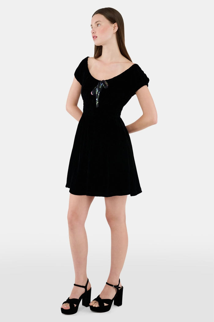 A woman faces forward in the Roller Rabbit Velvet Tilly Dress, a short-sleeve black velvet mini with a floral bow at the neckline, paired with black platform sandals, set against a plain light background.