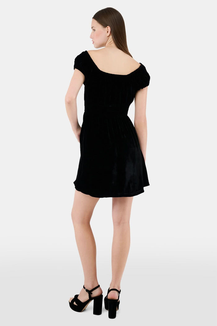 A woman faces forward in the Roller Rabbit Velvet Tilly Dress, a short-sleeve black velvet mini with a floral bow at the neckline, paired with black platform sandals, set against a plain light background.