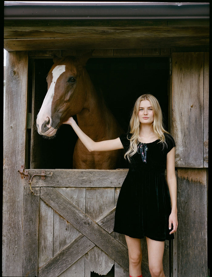 A woman faces forward in the Roller Rabbit Velvet Tilly Dress, a short-sleeve black velvet mini with a floral bow at the neckline, paired with black platform sandals, set against a plain light background.