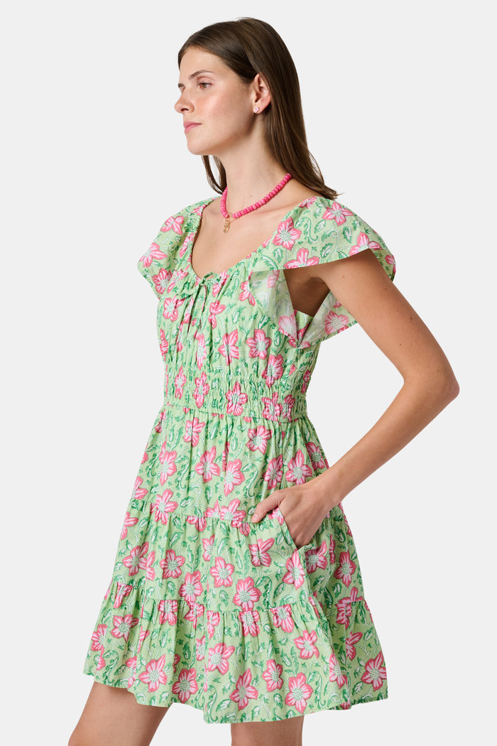 The Kids Verdant Floral Ryan Dress by Roller Rabbit is a green dress for little ones, showcasing pink and white hibiscus flowers in a floral block print. It features ruffled sleeves, a matching diaper cover, and is crafted from soft cotton for comfortable pullover wear.