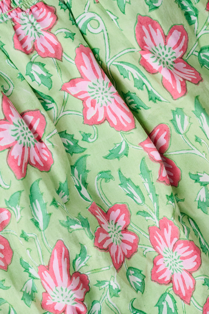The Verdant Floral Saylor Set by Roller Rabbit is a cotton two-piece girls’ outfit featuring a green background with a pink floral print. The set includes a ruffled, sleeveless top and matching shorts with an elastic waistband and ruffled hems.