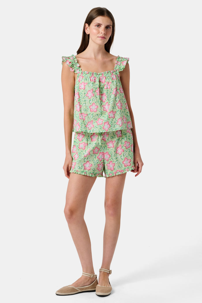 Three women are in a library. One wears the Verdant Floral Pipette Dress by Roller Rabbit—a green floral minidress—and stands with her hand on her chin. Another sits on a patterned sofa with her eyes closed, and bookshelves are visible in the background.