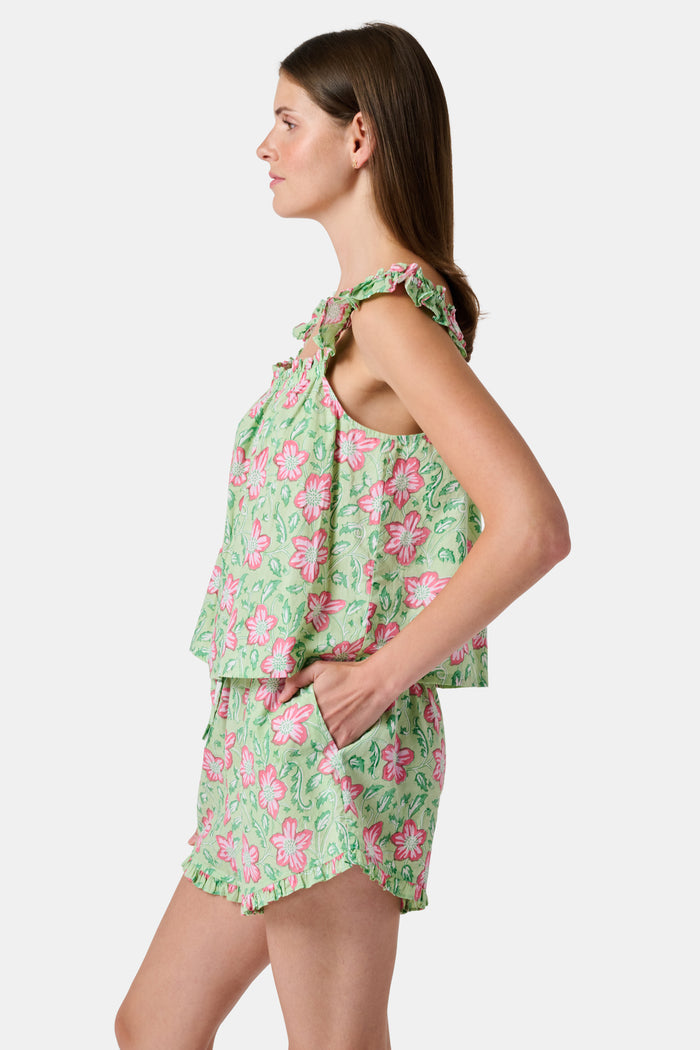 Three women are in a library. One wears the Verdant Floral Pipette Dress by Roller Rabbit—a green floral minidress—and stands with her hand on her chin. Another sits on a patterned sofa with her eyes closed, and bookshelves are visible in the background.