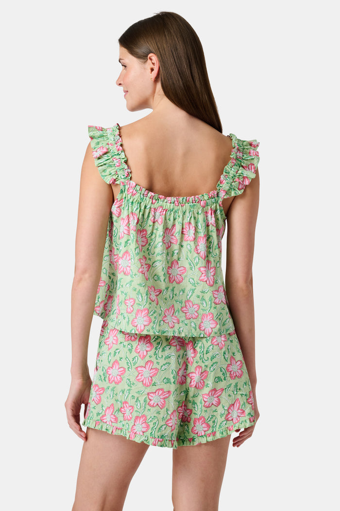 Three women are in a library. One wears the Verdant Floral Pipette Dress by Roller Rabbit—a green floral minidress—and stands with her hand on her chin. Another sits on a patterned sofa with her eyes closed, and bookshelves are visible in the background.