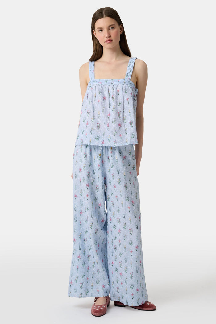 A woman with long brown hair wears the Roller Rabbit Wildflower Jemina Victoria Top, a sleeveless baby blue linen piece with wide shoulder straps, standing against a plain light background.