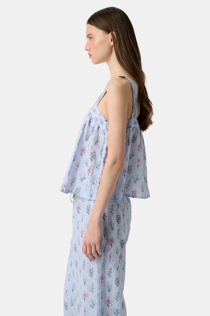 A woman with long brown hair wears the Roller Rabbit Wildflower Jemina Victoria Top, a sleeveless baby blue linen piece with wide shoulder straps, standing against a plain light background.