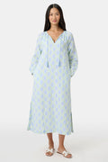 A woman faces forward in the Roller Rabbit Zelda Floral Shiv Caftan, a loose, ankle-length dress with a blue and green geometric print. She has long dark hair, white sandals, and stands against a plain white background.