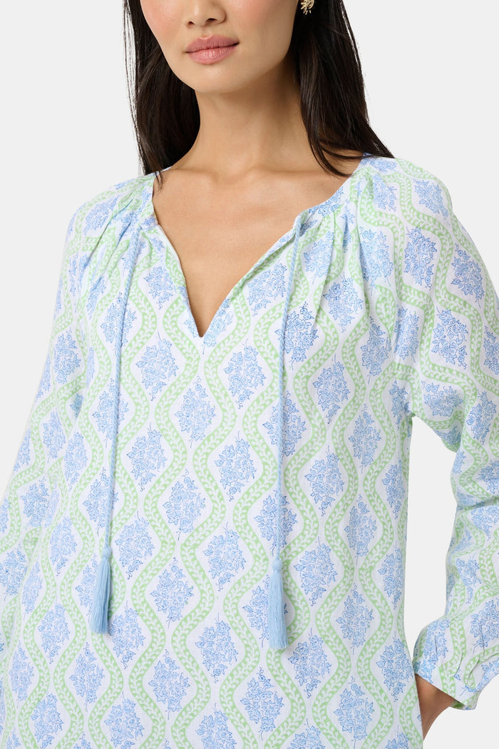 A woman faces forward in the Roller Rabbit Zelda Floral Shiv Caftan, a loose, ankle-length dress with a blue and green geometric print. She has long dark hair, white sandals, and stands against a plain white background.