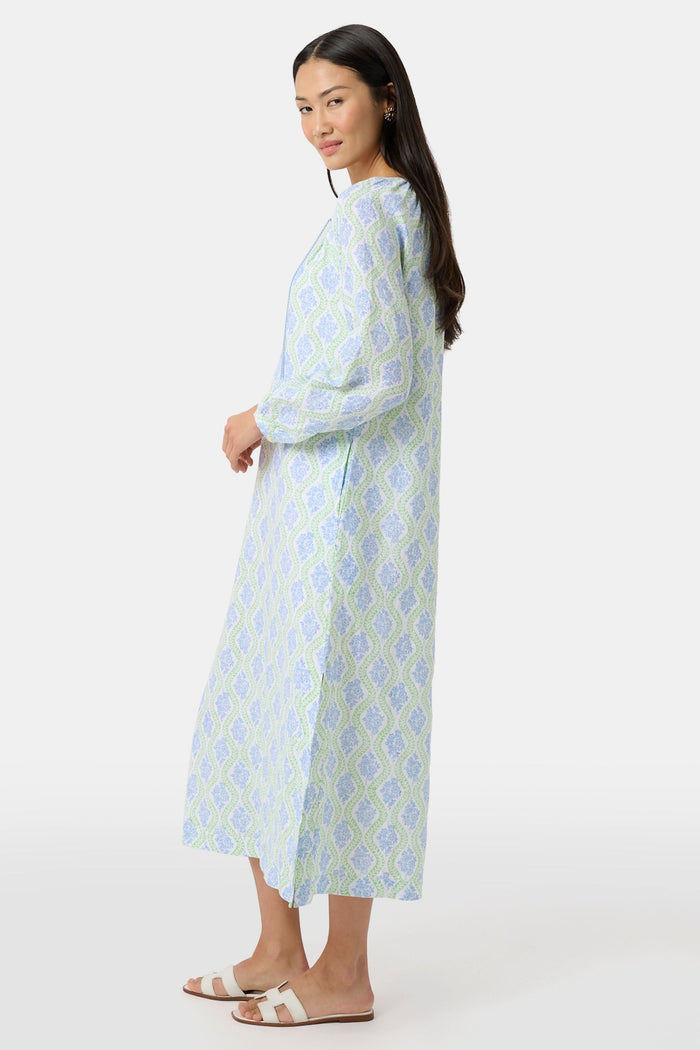 A woman faces forward in the Roller Rabbit Zelda Floral Shiv Caftan, a loose, ankle-length dress with a blue and green geometric print. She has long dark hair, white sandals, and stands against a plain white background.