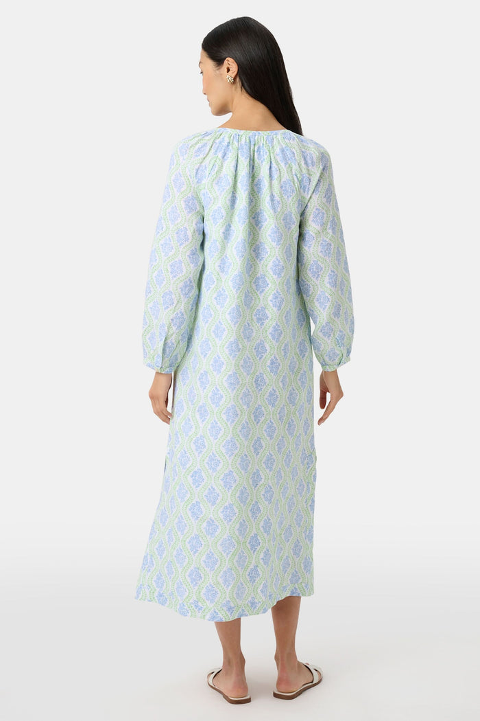 A woman faces forward in the Roller Rabbit Zelda Floral Shiv Caftan, a loose, ankle-length dress with a blue and green geometric print. She has long dark hair, white sandals, and stands against a plain white background.