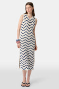 A woman wears the Roller Rabbit Zig Zag Irene Cover Up, a sleeveless, ankle-length crochet maxi dress with a blue and white zig zag pattern. She pairs it with black sandals and chunky striped bracelets, her straight brown hair worn loose.