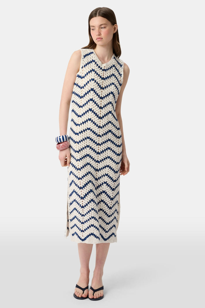 A woman wears the Roller Rabbit Zig Zag Irene Cover Up, a sleeveless, ankle-length crochet maxi dress with a blue and white zig zag pattern. She pairs it with black sandals and chunky striped bracelets, her straight brown hair worn loose.