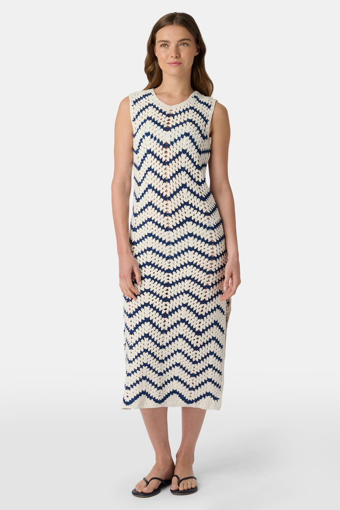 A woman wears the Roller Rabbit Zig Zag Irene Cover Up, a sleeveless, ankle-length crochet maxi dress with a blue and white zig zag pattern. She pairs it with black sandals and chunky striped bracelets, her straight brown hair worn loose.