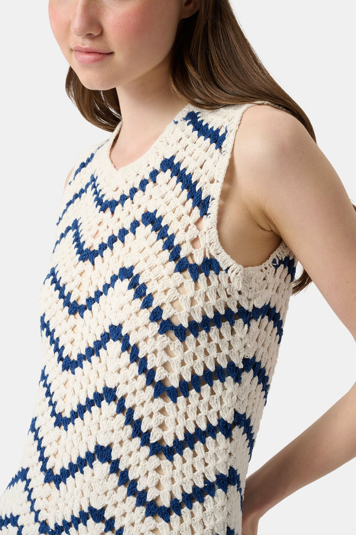 A woman wears the Roller Rabbit Zig Zag Irene Cover Up, a sleeveless, ankle-length crochet maxi dress with a blue and white zig zag pattern. She pairs it with black sandals and chunky striped bracelets, her straight brown hair worn loose.