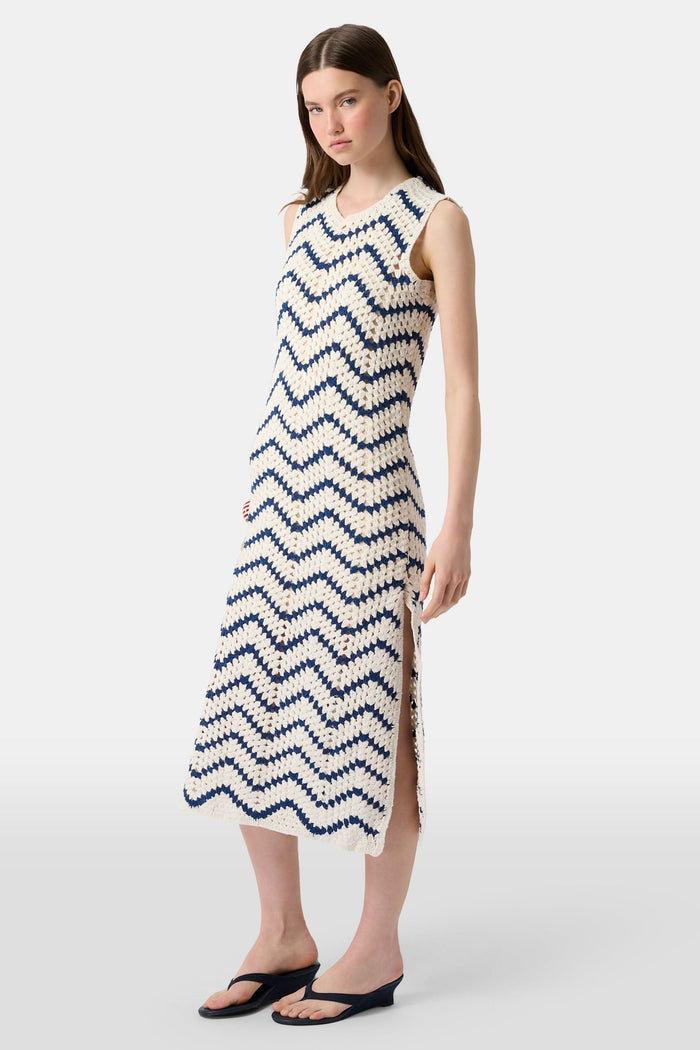 A woman wears the Roller Rabbit Zig Zag Irene Cover Up, a sleeveless, ankle-length crochet maxi dress with a blue and white zig zag pattern. She pairs it with black sandals and chunky striped bracelets, her straight brown hair worn loose.