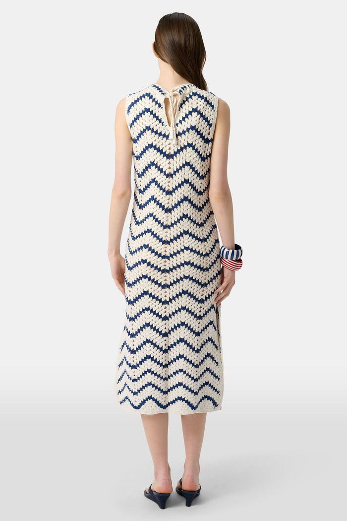 A woman wears the Roller Rabbit Zig Zag Irene Cover Up, a sleeveless, ankle-length crochet maxi dress with a blue and white zig zag pattern. She pairs it with black sandals and chunky striped bracelets, her straight brown hair worn loose.