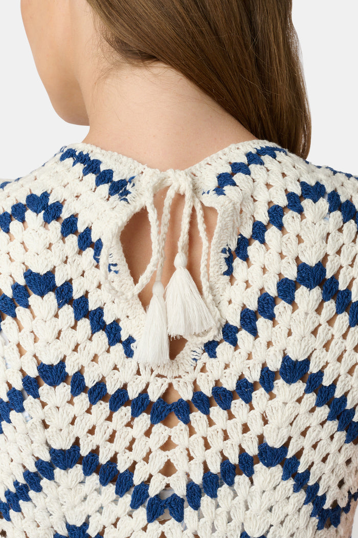 A woman wears the Roller Rabbit Zig Zag Irene Cover Up, a sleeveless, ankle-length crochet maxi dress with a blue and white zig zag pattern. She pairs it with black sandals and chunky striped bracelets, her straight brown hair worn loose.