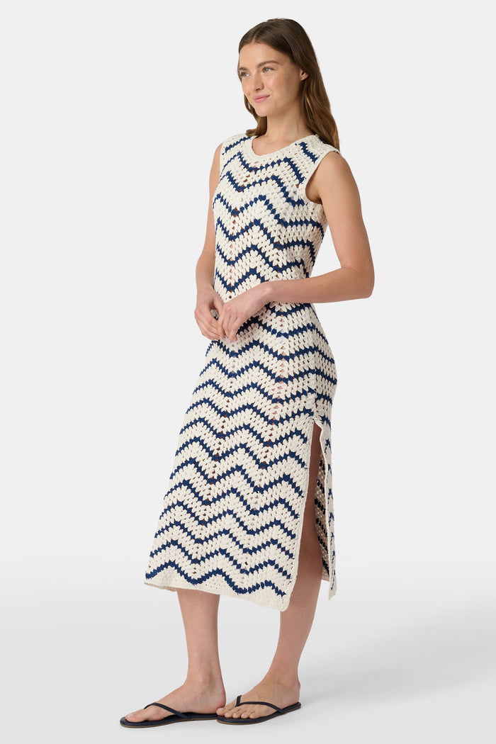 A woman wears the Roller Rabbit Zig Zag Irene Cover Up, a sleeveless, ankle-length crochet maxi dress with a blue and white zig zag pattern. She pairs it with black sandals and chunky striped bracelets, her straight brown hair worn loose.