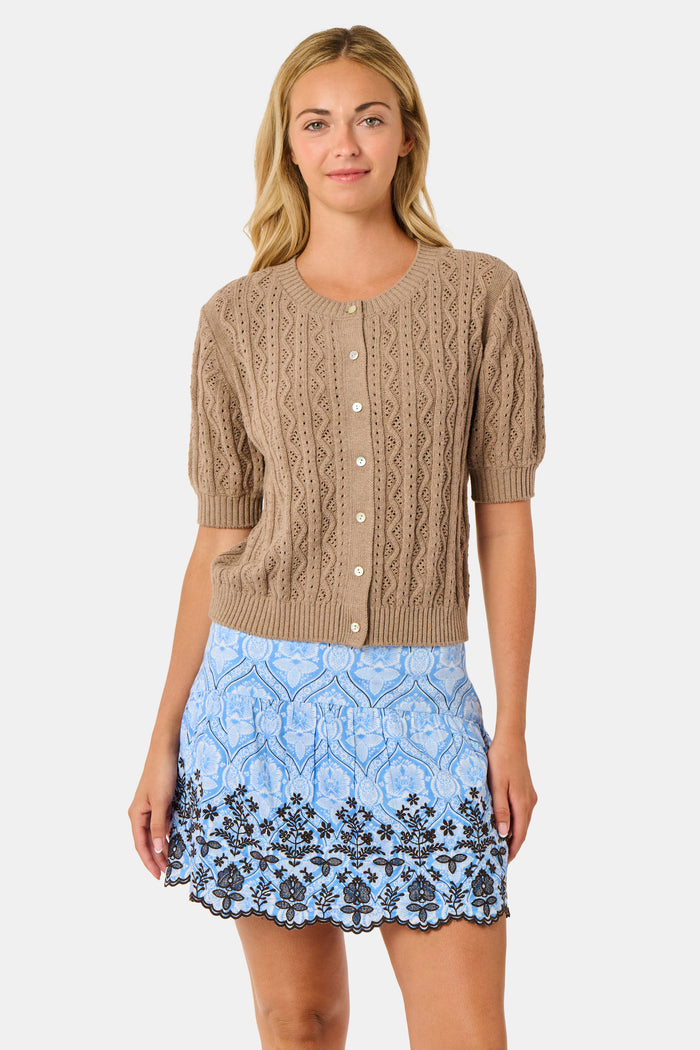 A woman wears the Roller Rabbit Zig Zag Pointelle Nyla Cardigan, a short-sleeved, walnut brown button-front sweater with circular patterns, paired with a light blue patterned skirt.