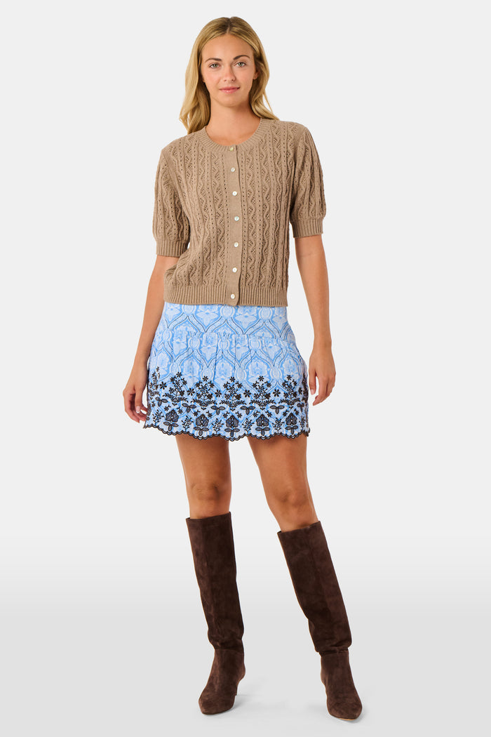 A woman wears the Roller Rabbit Zig Zag Pointelle Nyla Cardigan, a short-sleeved, walnut brown button-front sweater with circular patterns, paired with a light blue patterned skirt.