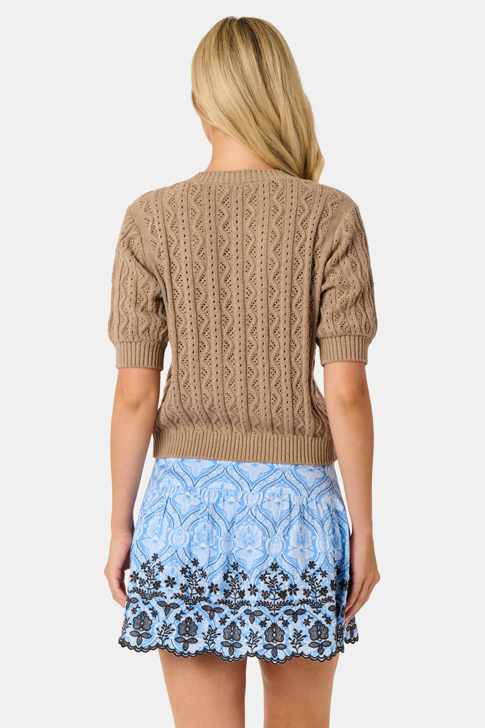 A woman wears the Roller Rabbit Zig Zag Pointelle Nyla Cardigan, a short-sleeved, walnut brown button-front sweater with circular patterns, paired with a light blue patterned skirt.
